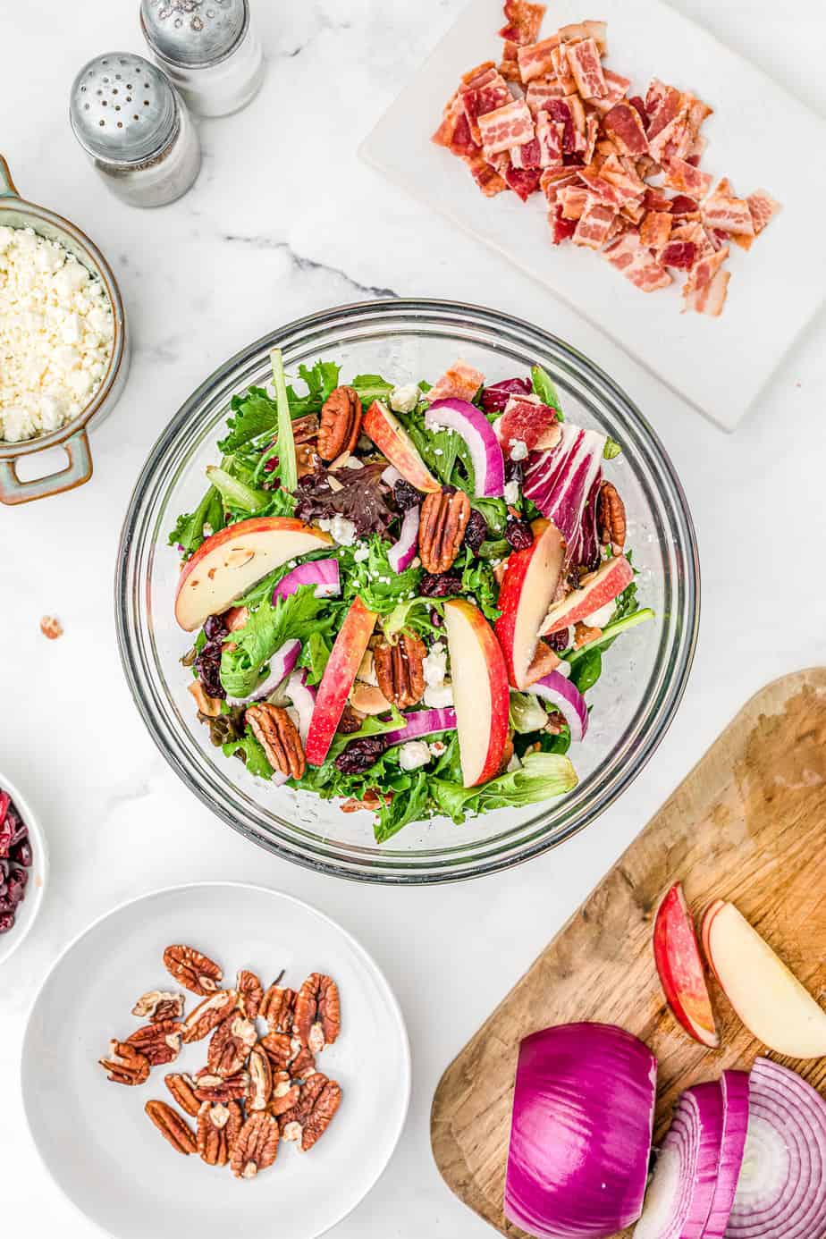 Sliced apples, pecans, cranberries, slices of onion and chopped bacon being added to a large bowl full of spring mix salad on a counter from overhead with more  of the salas ingredients in bowls nearby.