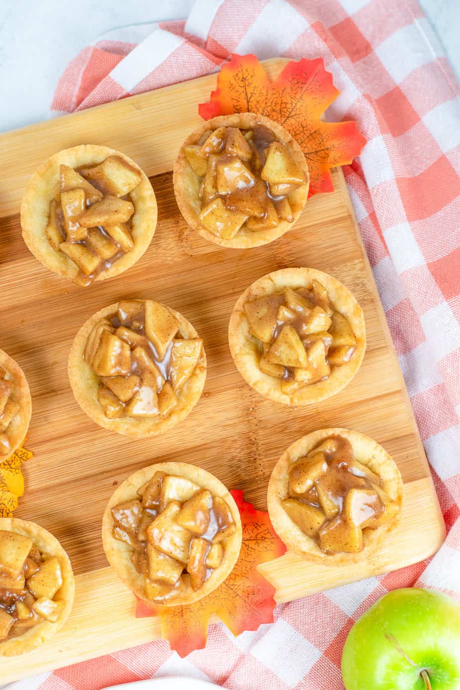 Three rows of apple crisp cookie cups lined up on a wood platter from overhead with a napkin and an apple nearby on the kitchen counter.