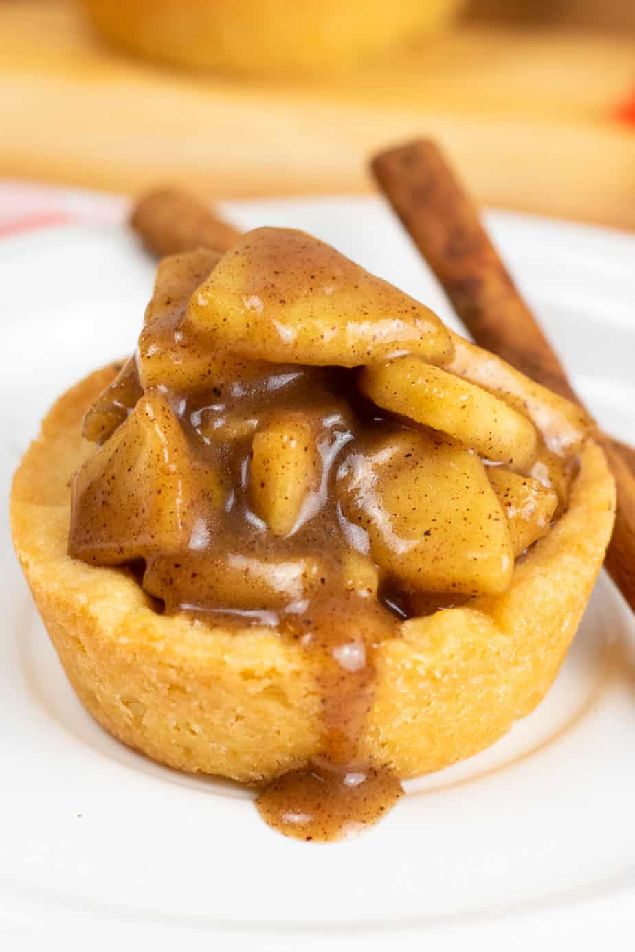 Very close up side view of a sugar cookie cup full of gooey apple filling drizzling out the side on a plate next to a cinnamon stick.