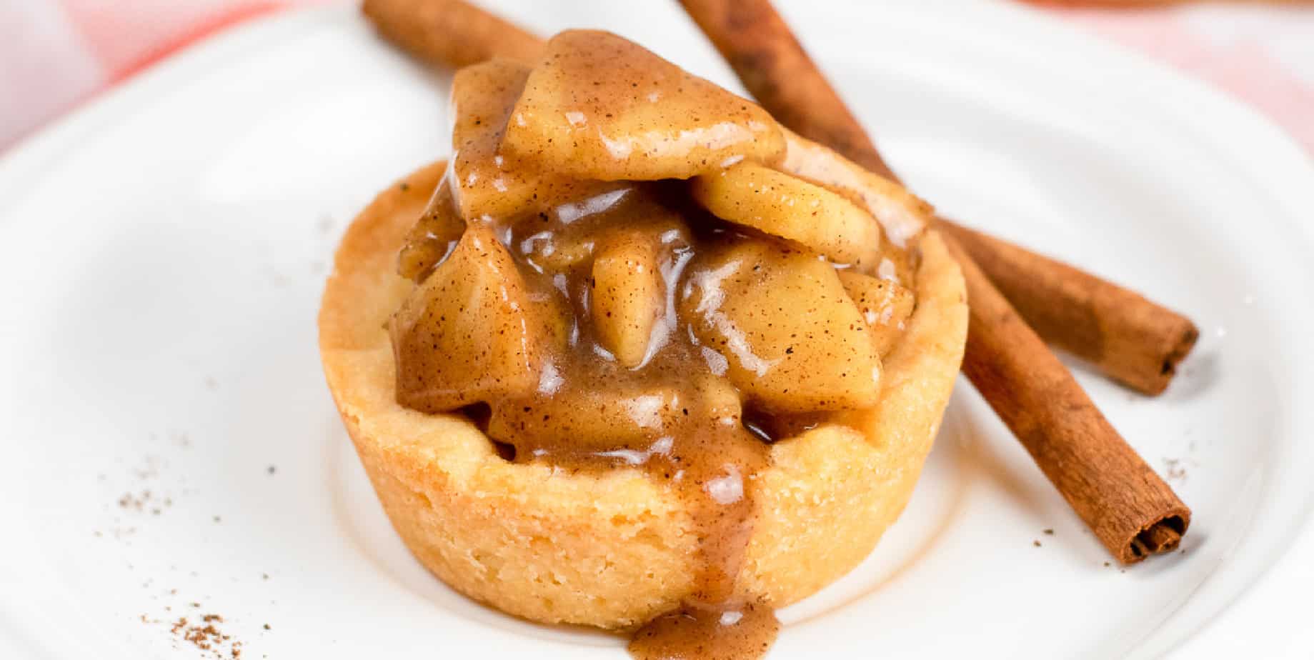 Wide close up side view of an apple crisp cookie cup full of gooey apples on a plate from the side next to sticks of cinnamon.