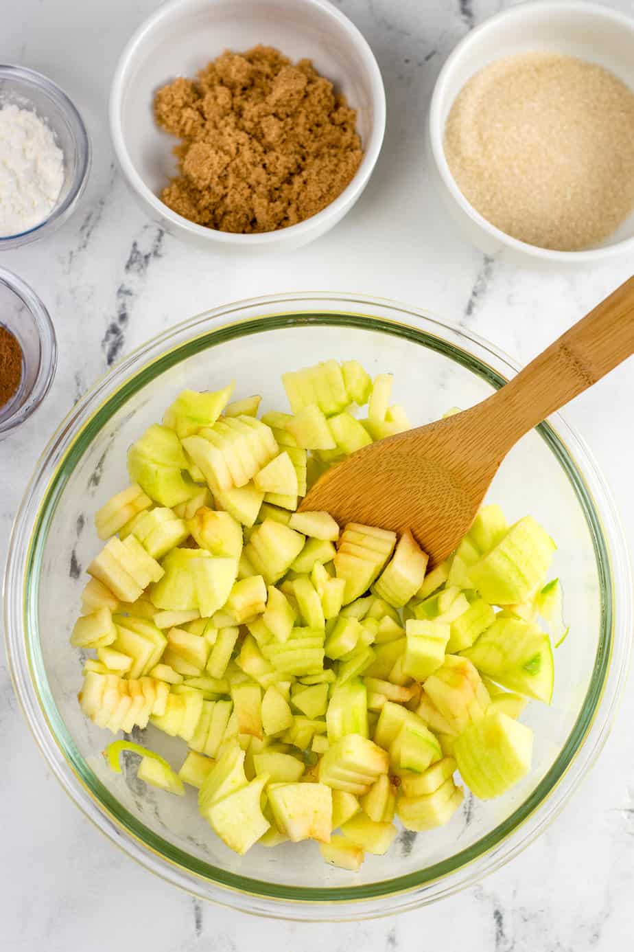 Apple cut up into small chunks place in a large mixing bowl with a spatula on a counter from overhead with bowls of cinnamon, sugar and cornstarch nearby.