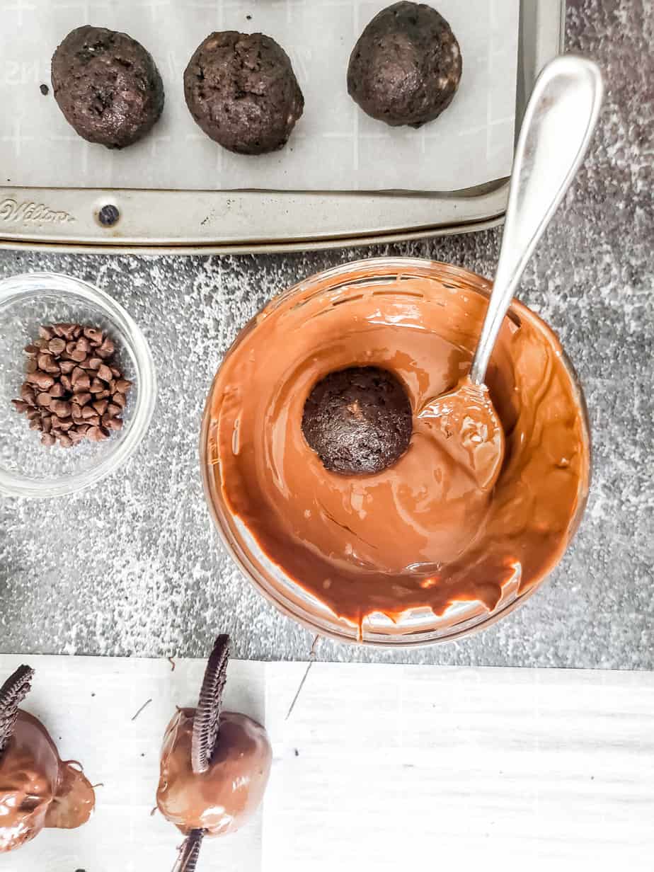 Chocolate oreo balls being dipped in a bowl of melted chocolate with some truffles on a pan and some with oreo bat wings looking from above.