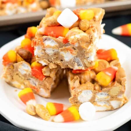 Side view of three candy corn peanut butter bars stacked on a plate with more candy corn, marshmallows and peanuts nearby on the plate close up.