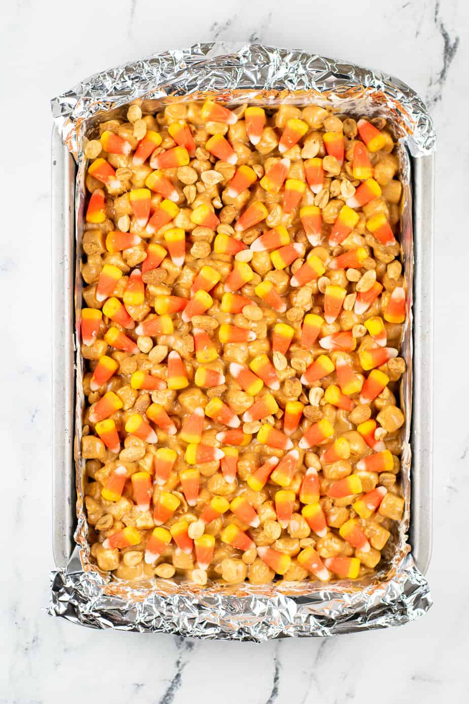 Large rectangular pan from above lined with aluminum foil and full of peanut butter marshmallow bars topped with more candy corn and peanuts.