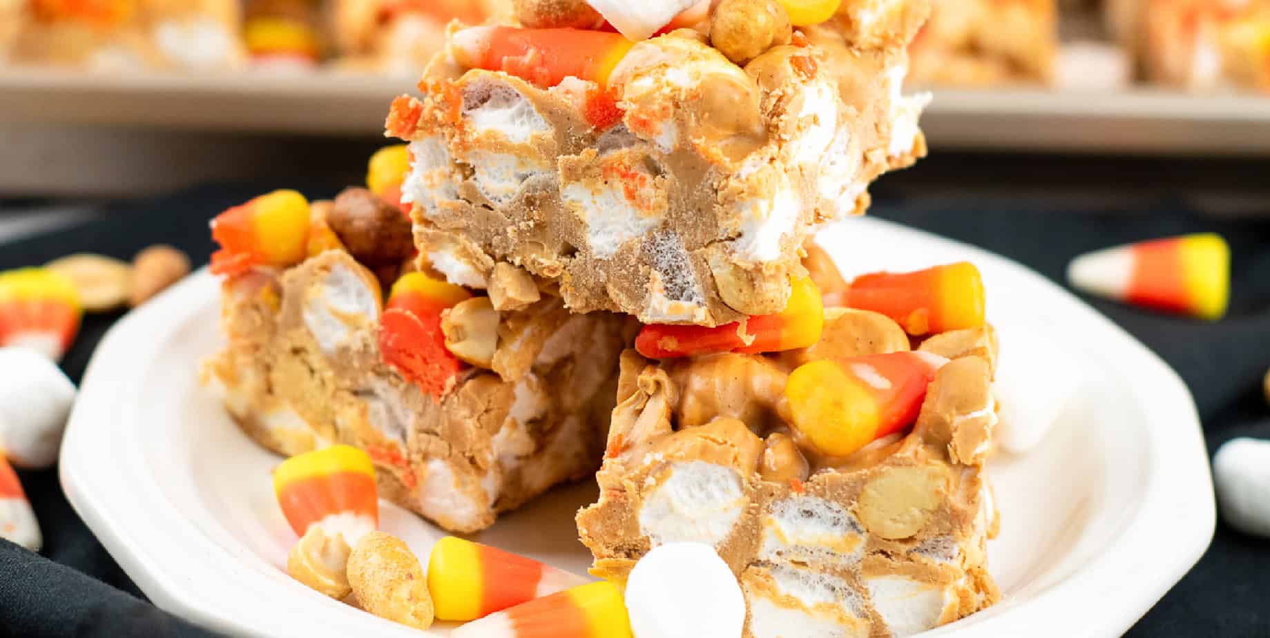 Wide view from the side of candy corn peanut butter marshmallow bars from the side on a plate on a black counter.