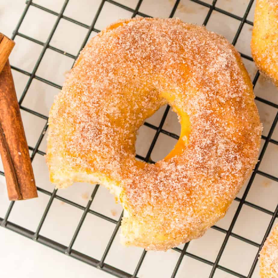 Close up overhead of a cinnamon sugar coated donut with a bite missing cooling on a wire rack with a stick of cinnamon nearby on the counter.