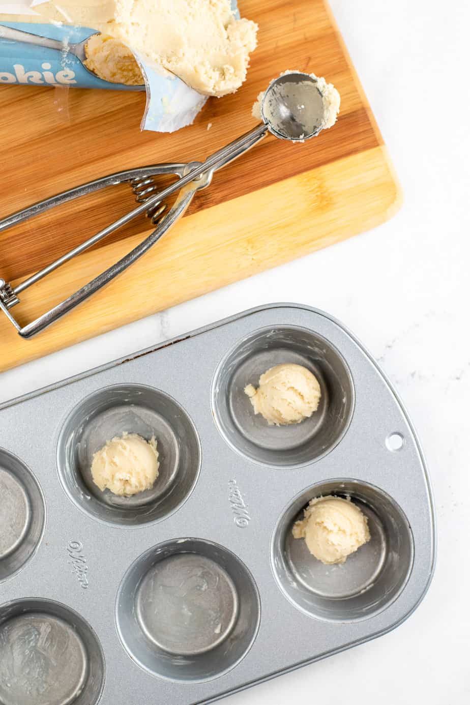 Cookie dough balls being scooped from a tube of cookie dough with a cookie scoop with a greased muffin tin with individual balls of cookie dough in each muffin well on a kitchen counter from overhead.