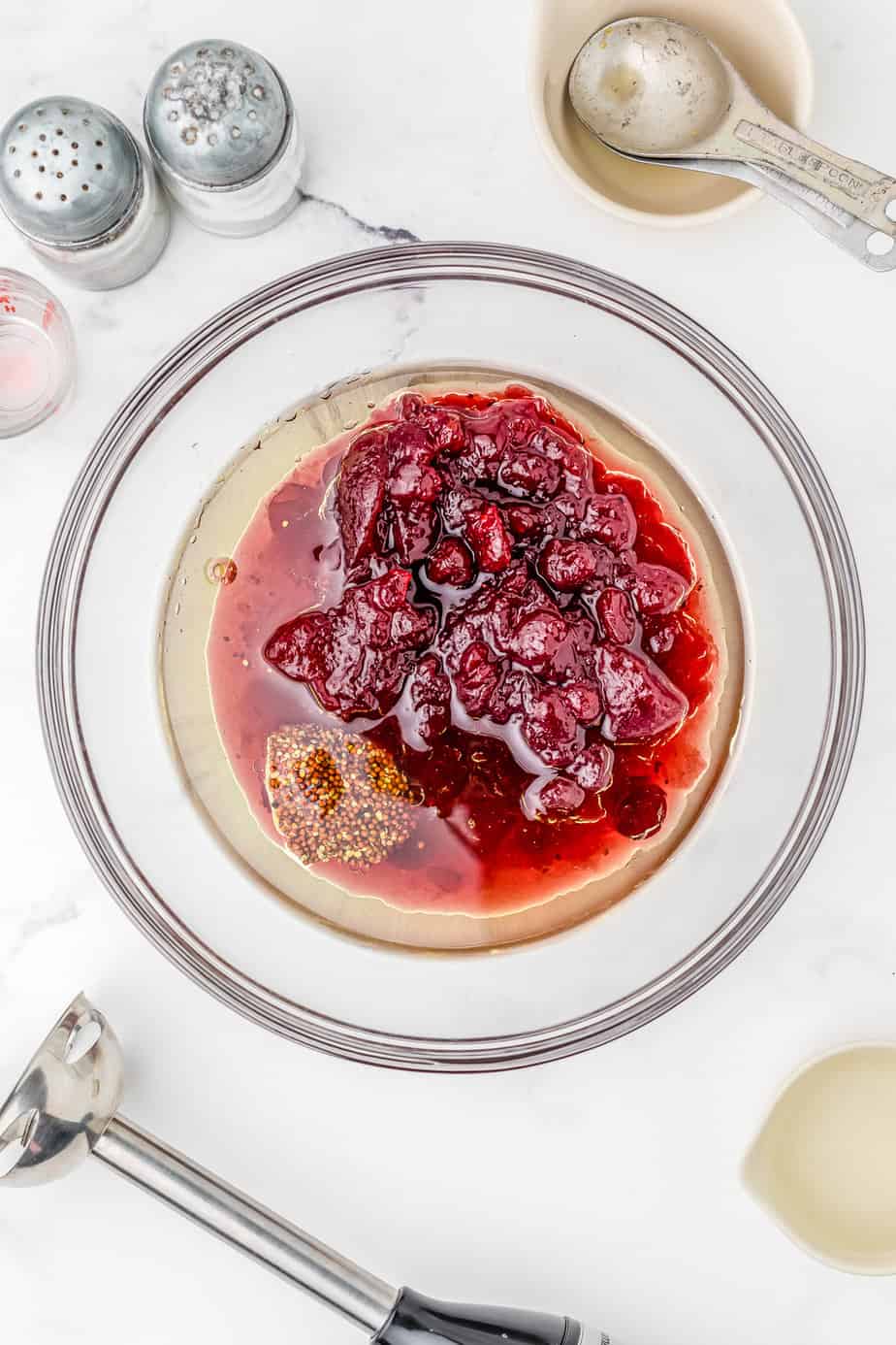Cranberry sauce, oil, mustard and maple syrup in a bowl on a counter from overhead with an immersion blender, measuring cups and salt and pepper also on the counter.
