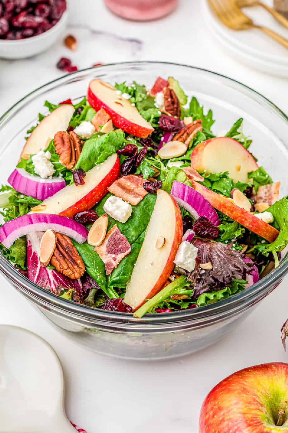 Large bowl of salad from the side on a table topped with apple slices, dried cranberries, pecans, onions and feta cheese with serving utensils and extra ingredients next to the bowl on the table.