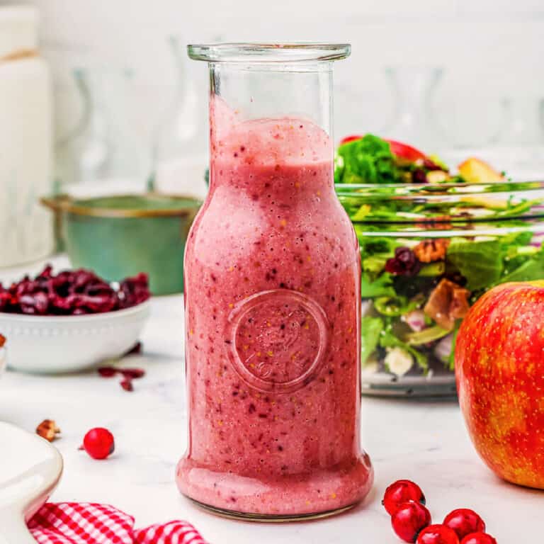 Cranberry Vinaigrette in a large glass jar from the side on a kitchen counter with salad in a glass bowl, dried cranberries in a bowl, and a fresh apple around the dressing.