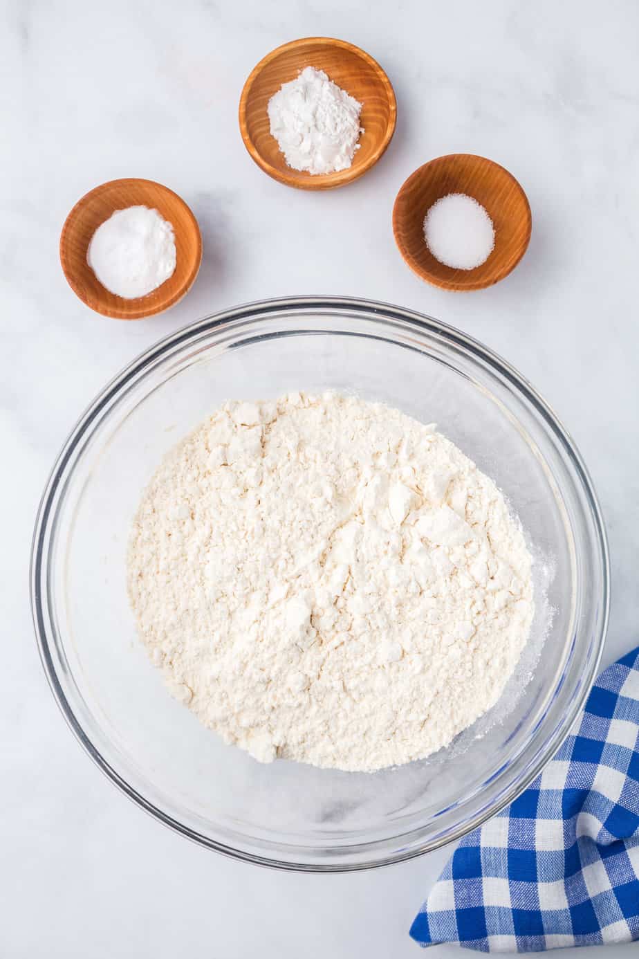 Flour in a large mixing bowl with other dry ingredients in small bowls nearby on a counter from overhead.