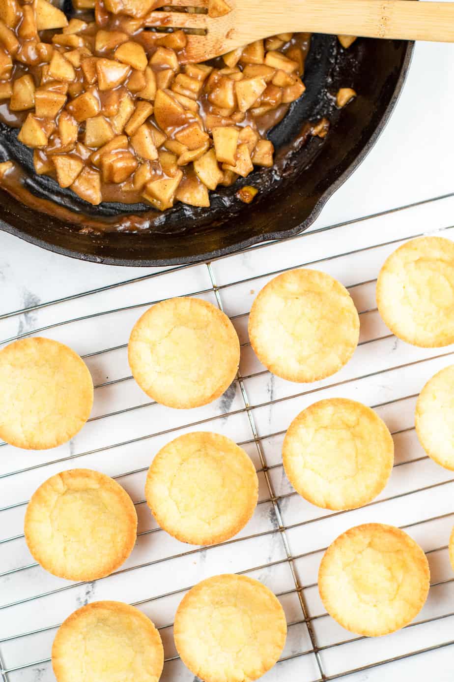 Empty sugar cookie cups on a wire rack next to a pan of cooked apple pieces from above on a counter.