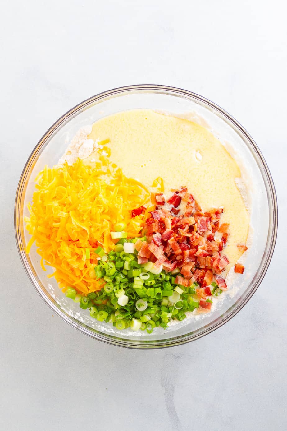 Shredded cheddar cheese, diced green onions and diced bacon on top of a yellow dough in a large mixing bowl from above on a counter.