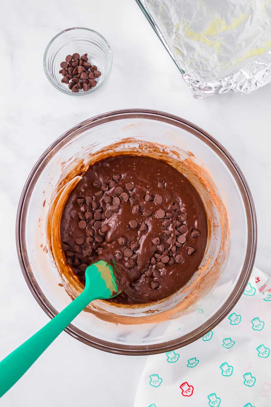 Folding chocolate chips into batter in a large bowl with a silicone spatula with a pan and a small bowl with a few chocolate chips on the counter from overhead.