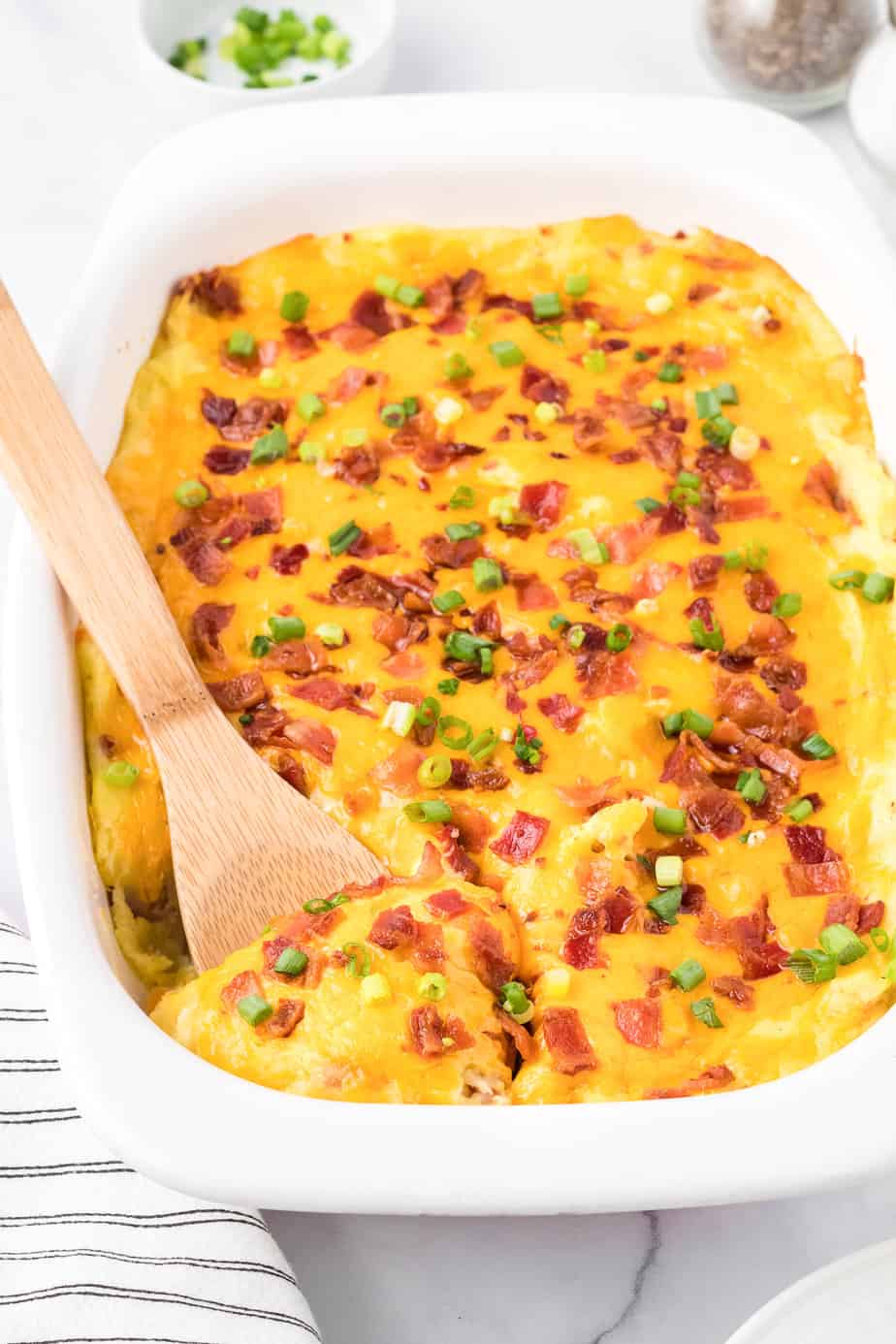 Side view of a pan of loaded mashed potato casserole on a counter topped with cheese and bacon being scooped with a wooden spoon.