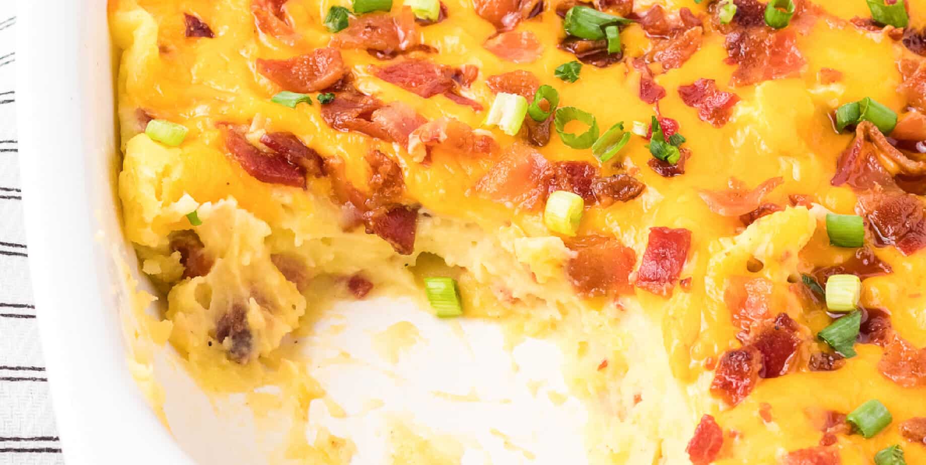 Close up on a pan of loaded mashed potato casserole missing a scoop covered in cheese and bacon from the side.