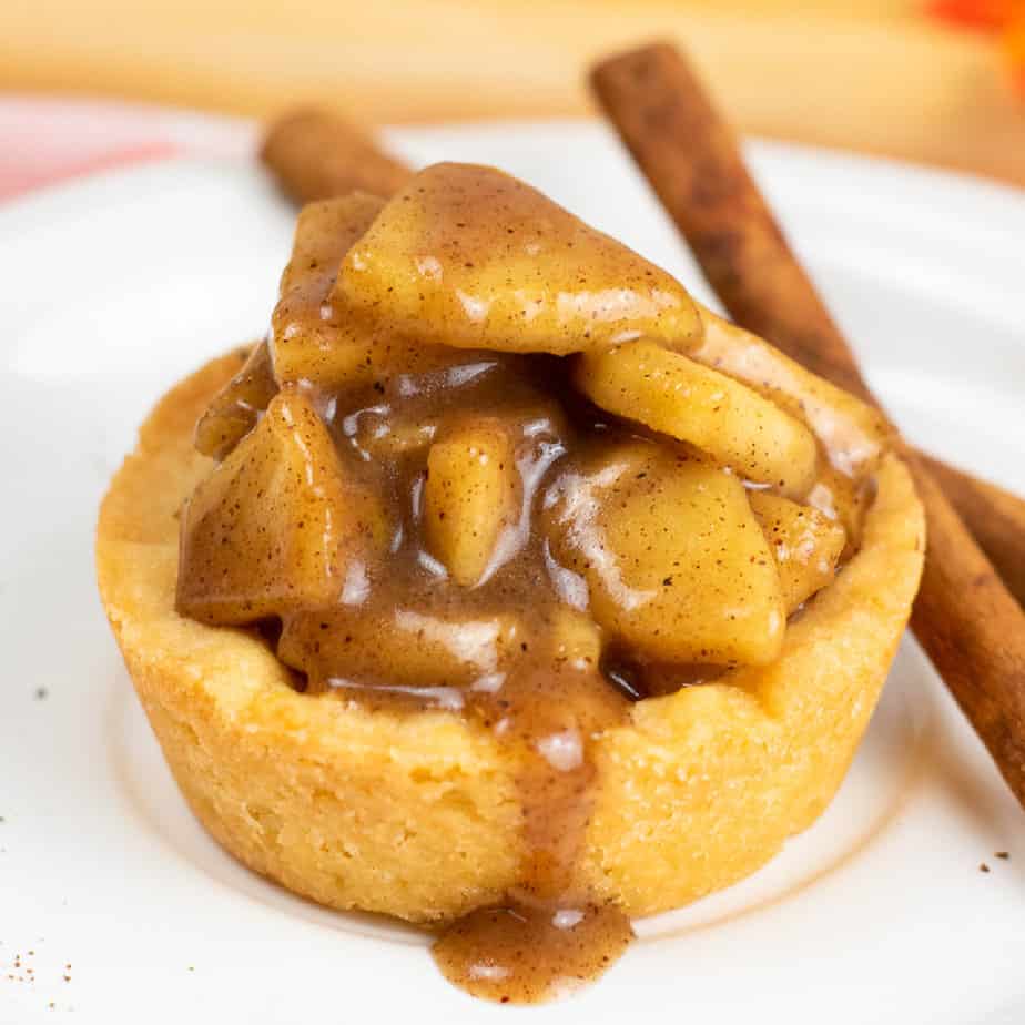 Close up side view of a sugar cookie cup filled with a gooey apple filling with some drizzling out on the plate below from the side next to a few sticks of cinnamon.