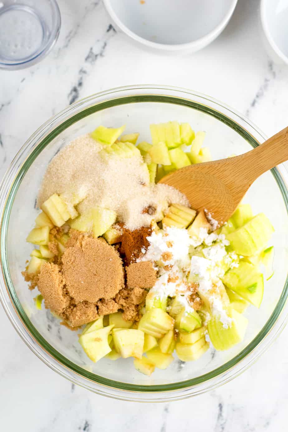 Mixing apples pieces with sugar, cinnamon and spices in a large mixing bowl on a counter from above.