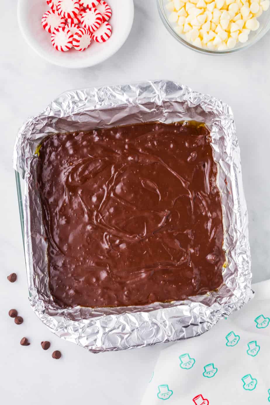 Raw brownie batter with chocolate chips in a square pan lined with aluminum foil from above with small bowls of peppermint candies and white chocolate chips on a counter from above.