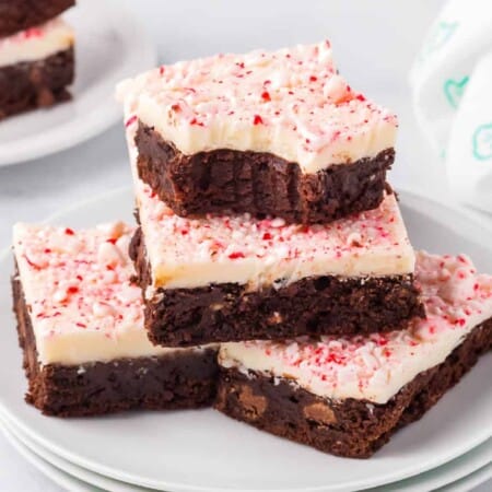 Brownies topped with white chocolate and pieces of peppermint stacked in a pile on a plate on a counter from the side with the top brownie missing a bite.