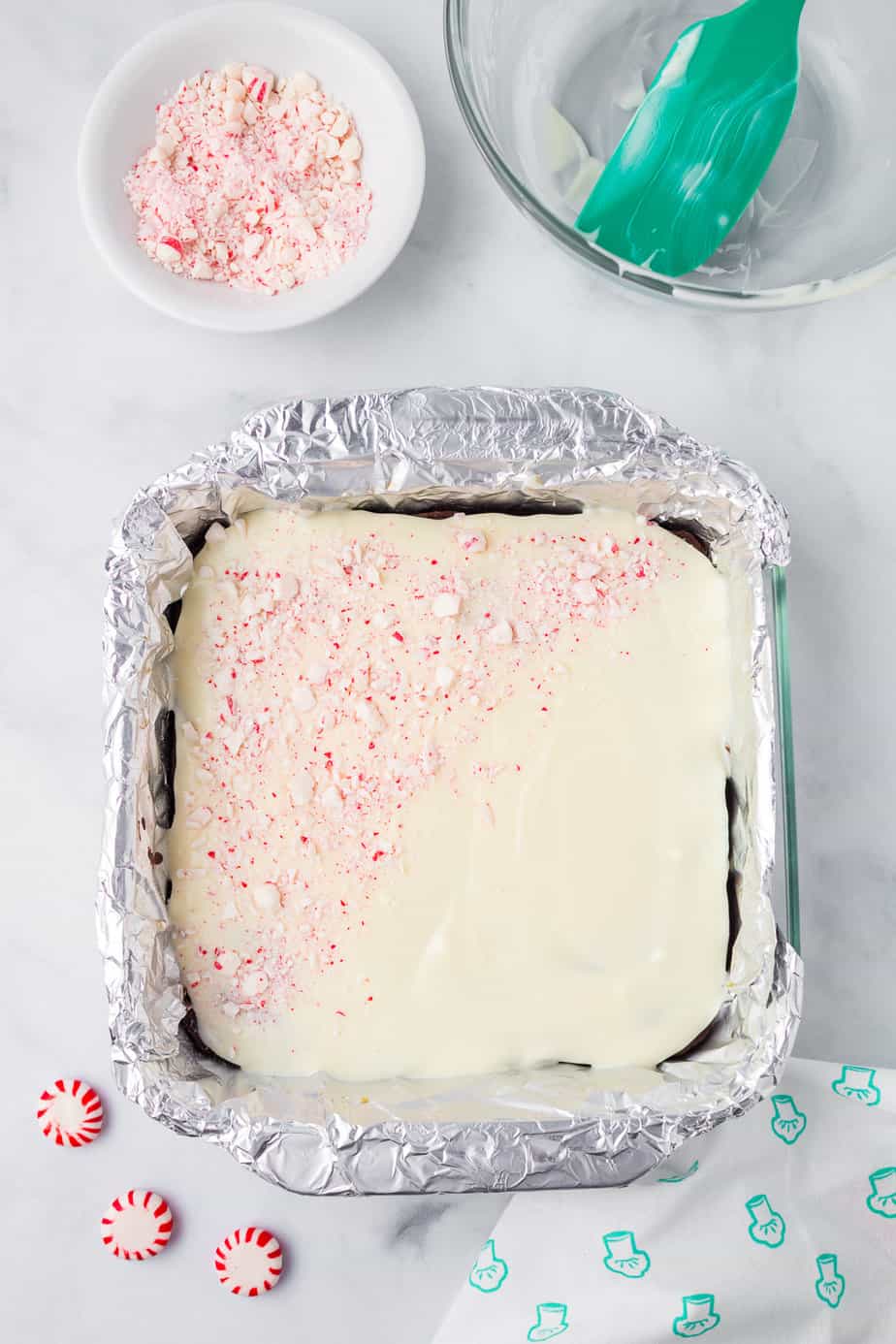 Brownies in the pan layered with melted white chocolate and broken pieces of peppermint candy from above with a bowl of more broken peppermint candies and a bowl with a spatula nearby on the counter.