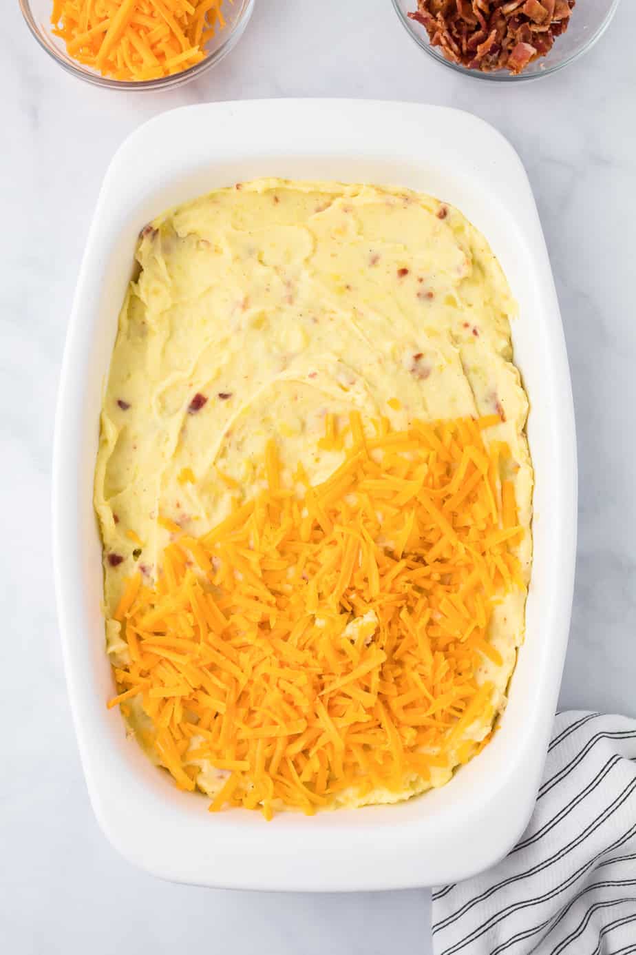 Adding cheese to a pan full of mashed potatoes in a rectangular casserole dish from above on a counter.