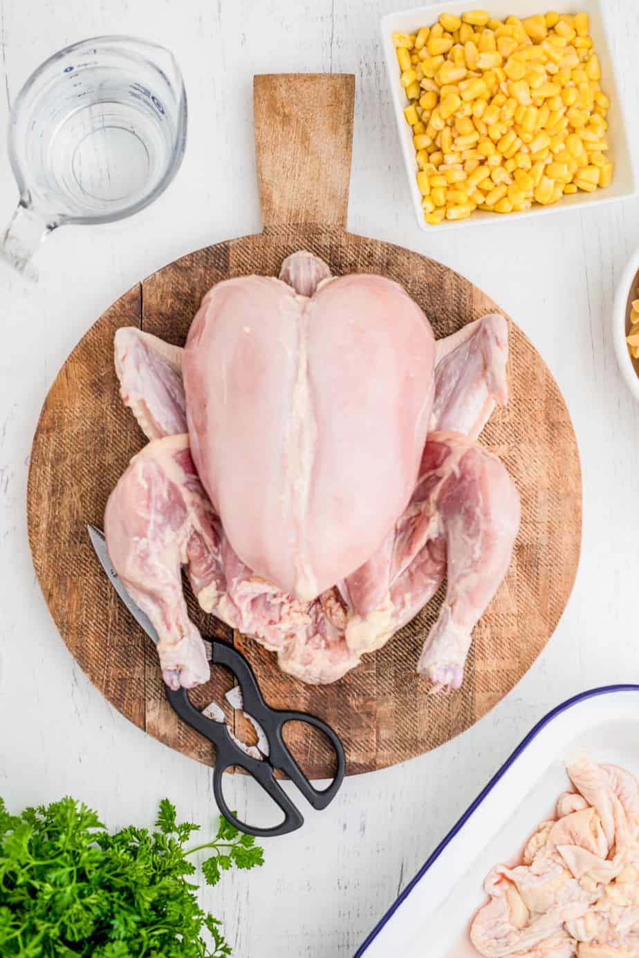 A whole raw chicken on a round cutting board with skin removed from above next to kitchen sheers, a pan full of chicken skin pieces, fresh parsley, corn in a bowl and water in a glass measuring cup on a counter from above.