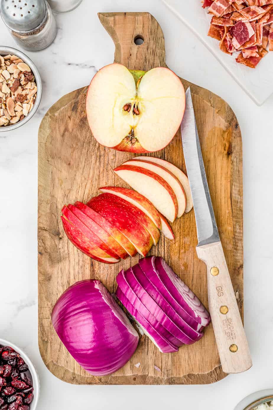 An apple half sliced and an onion all the way sliced into thin slices pn a cutting board from overhead on a counter next to a knife.