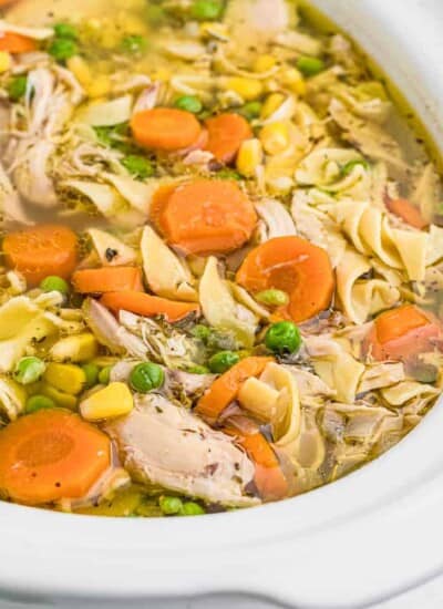 Up close view of an open slow cooker full of chicken, carrrots, peas, corn and noodles in a broth.