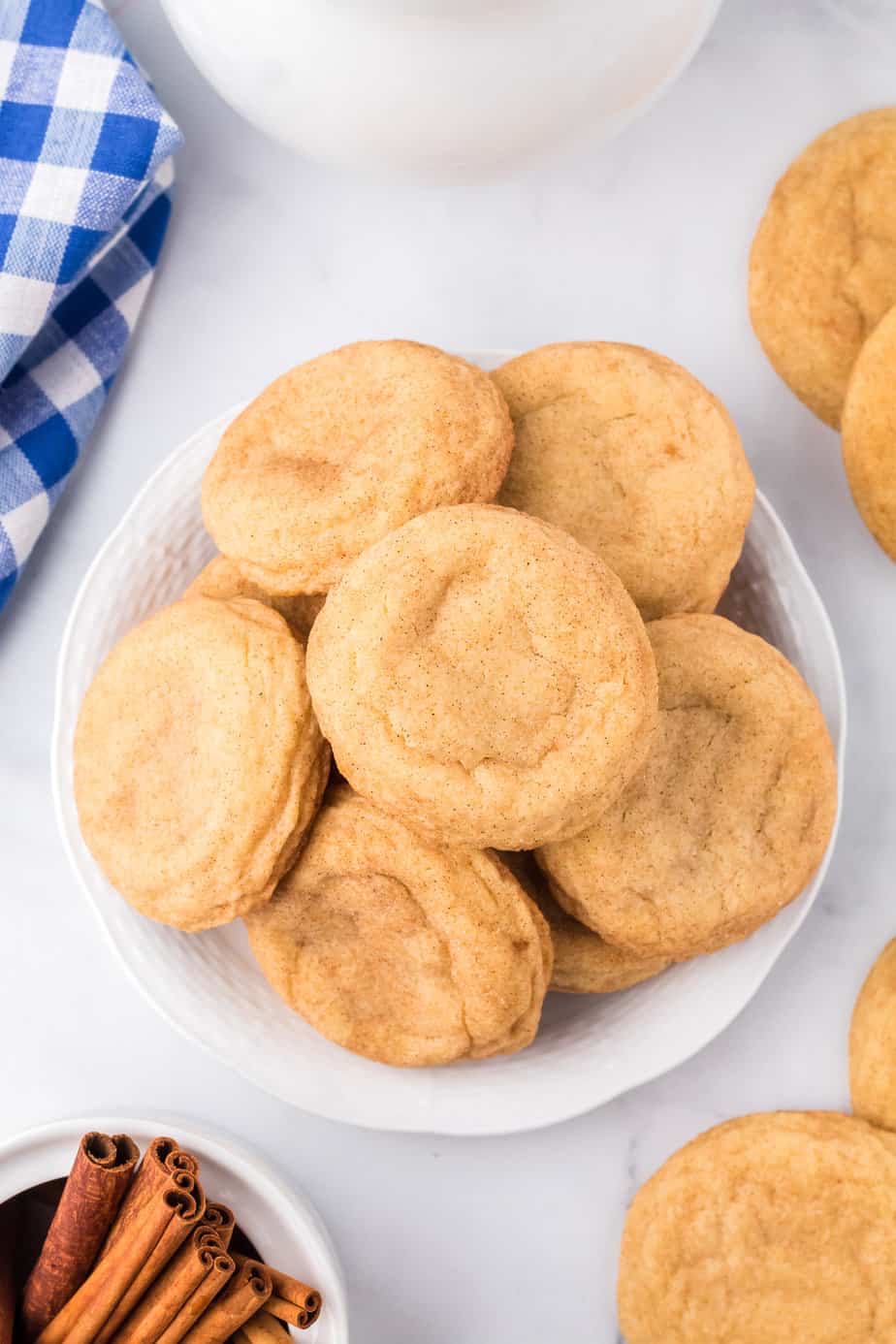 Snickerdoodle cookies on a plate piled high from overhead with cinnamon, more cookies, a napkin and milk on the counter nearby.