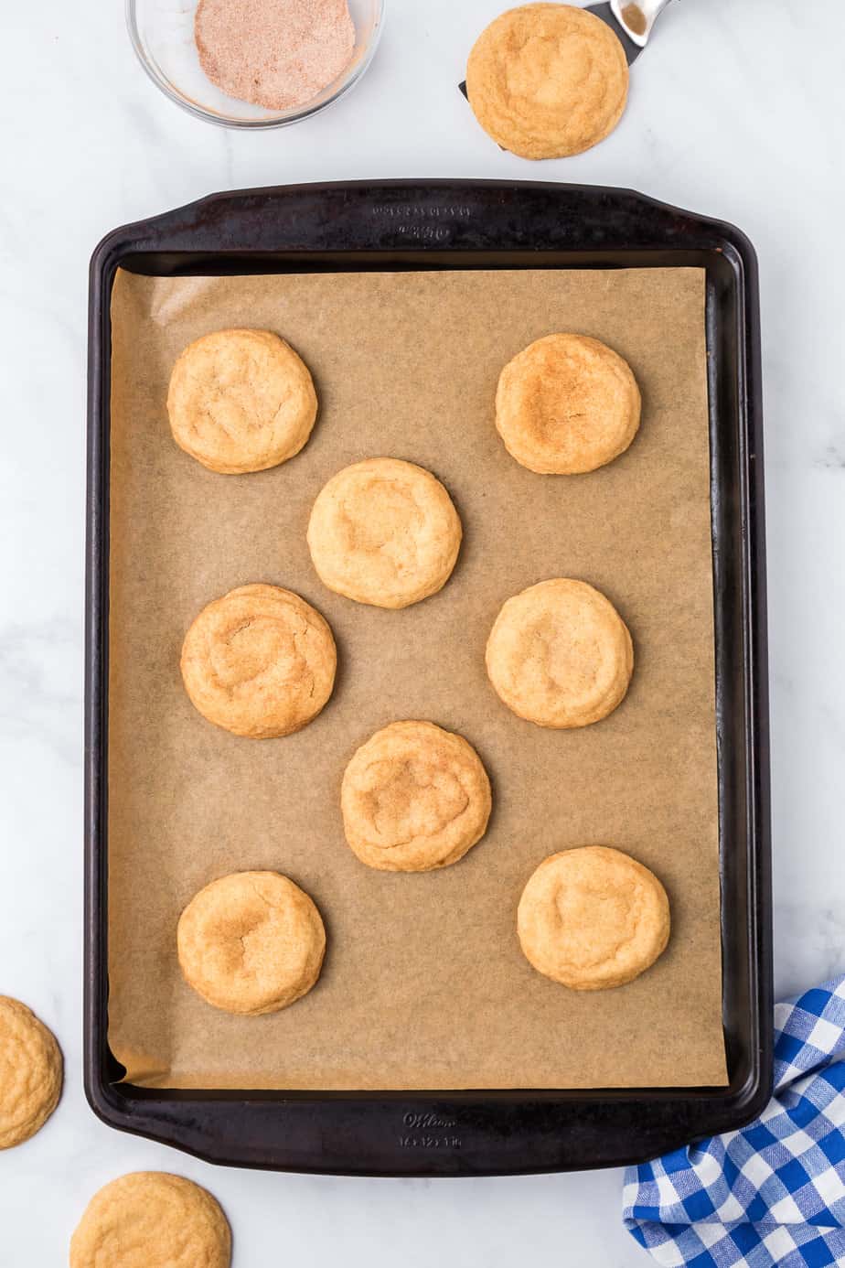 Snickerdoodle cookies baked on a pan lined with brown parchment on a counter from overhead with cinnamon sugar and a towel nearby.