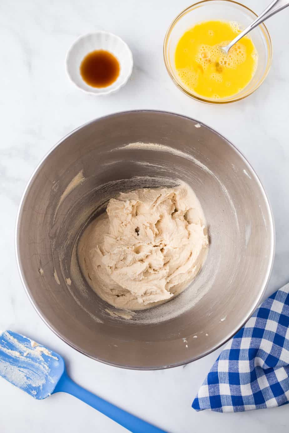 Butter and sugar creamed together in the bowl of a stand mixer from overhead on a counter with beaten egg and vanilla in bowls nearby on the table.