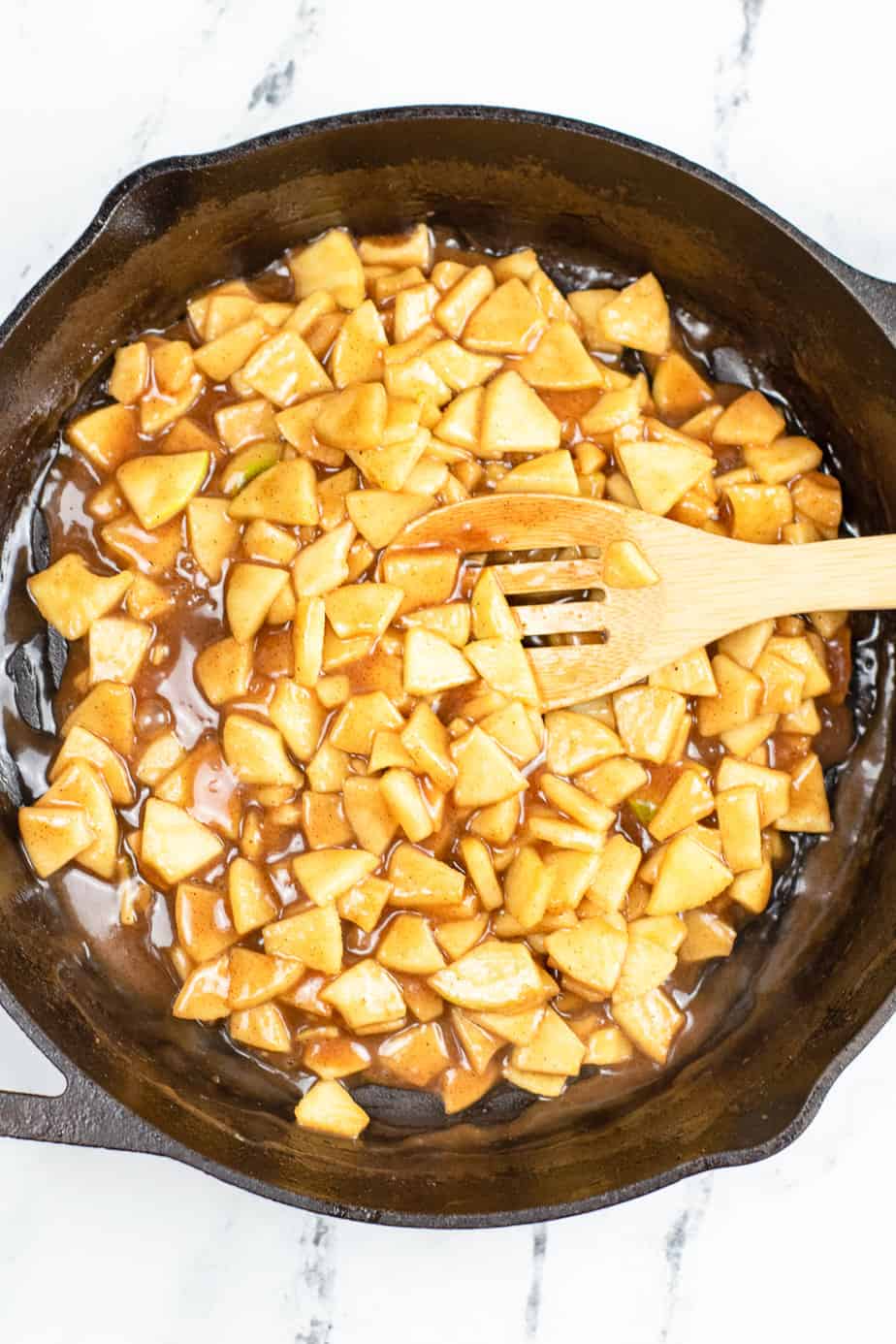 Cinnamon sugar seasoned apple pieces in a skillet with a spatula from overhead being cooked until softened and just beginning to caramelize.