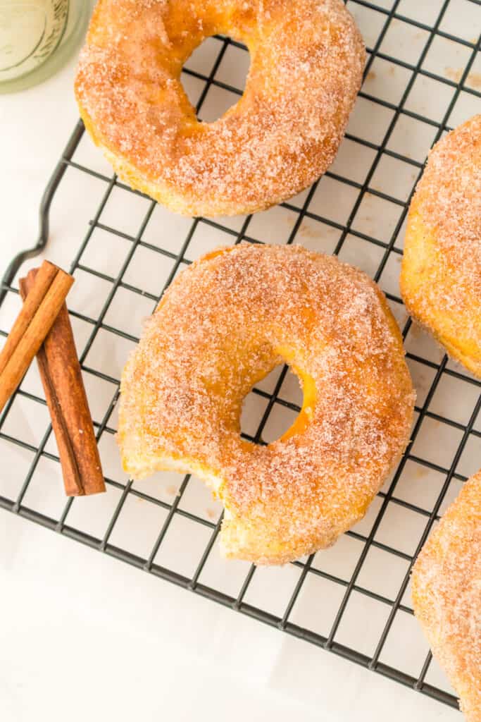 Three cinnamon sugar coated biscuit dough donuts cooling on a wire rack on a counter from above with one donut missing a bite.