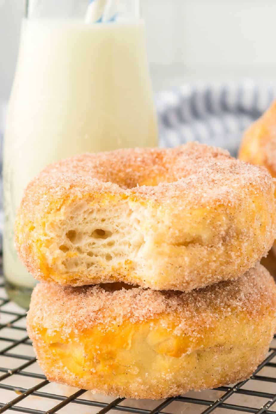 Two stacked donuts on a wire cooling rack on a counter from the side with the top donut missing a bite and a glass of milk behind.