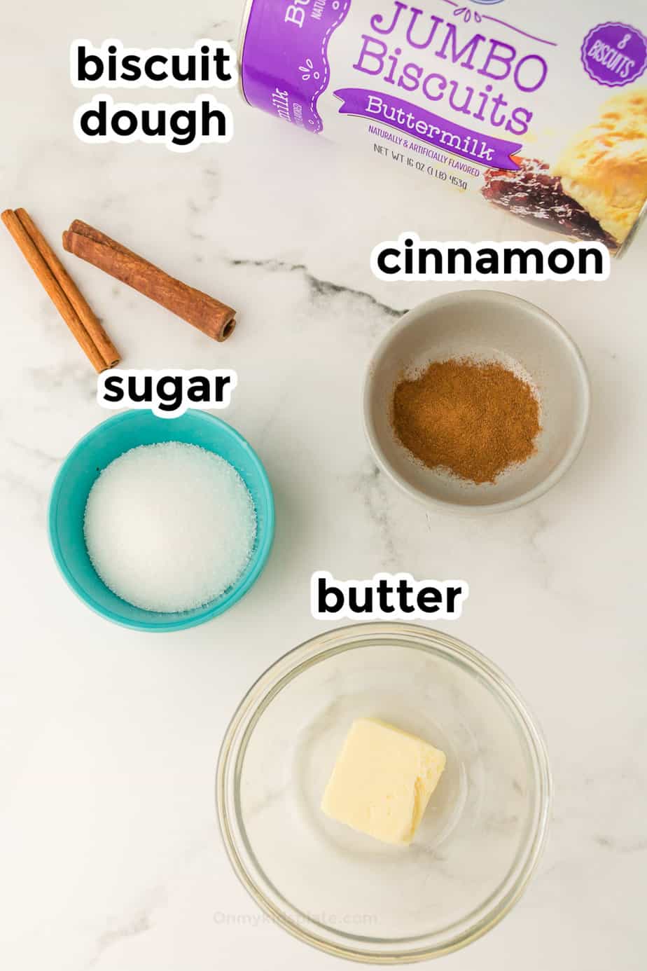 Cinnamon, butter, sugar and biscuit dough on a counter in bowls from overhead with labels.