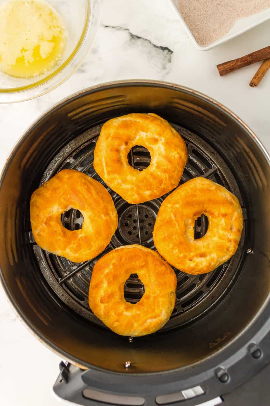 Four golden brown donuts made of biscuit dough in the basket of an air fryer from above on the counter with a bowl of melted butter and cinnamon sugar nearby on the counter.