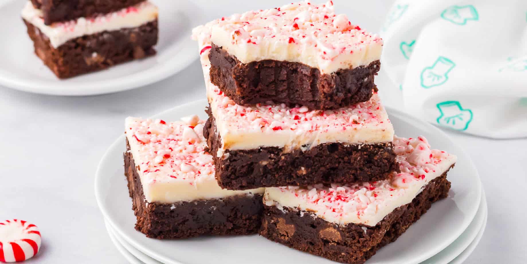 Brownies topped in white chocolate and peppermint piled on a plate on a counter with the top brownie missing a bite and more brownies on a plate in the background.