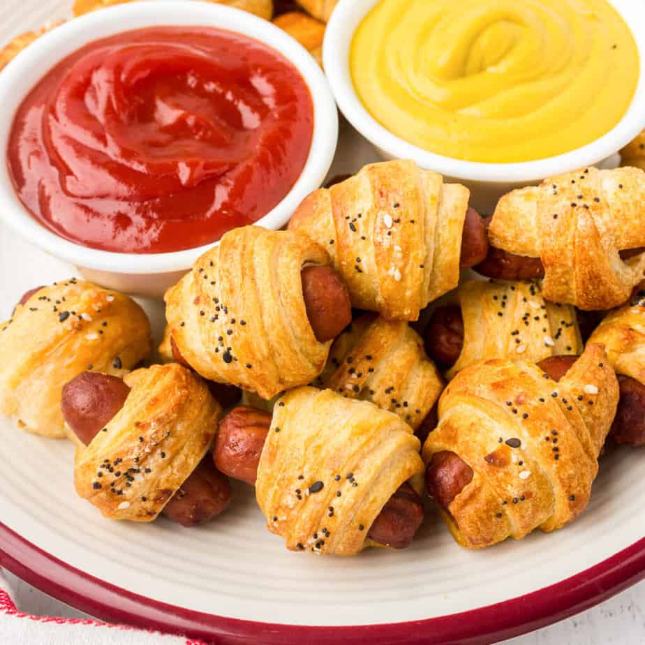 Pigs in a blanket on a plate from above with bowls of ketchup and mustard on the plate.