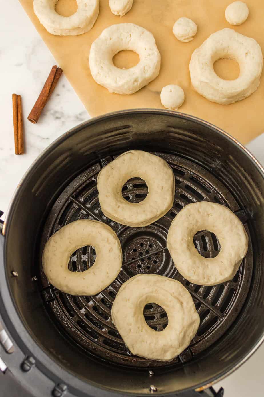 Four round donuts with centers missing placed in the basket of an air fryer from above on the counter with a cutting board and more extra raw doughnuts nearby.