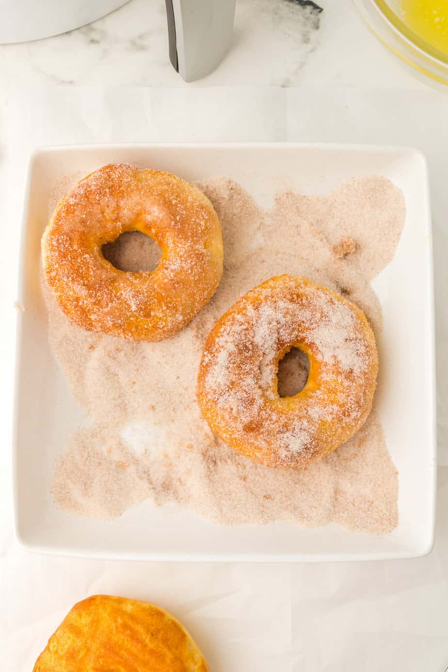 Two donuts being rolled in cinnamon sugar in a shallow square bowl from above on a counter.