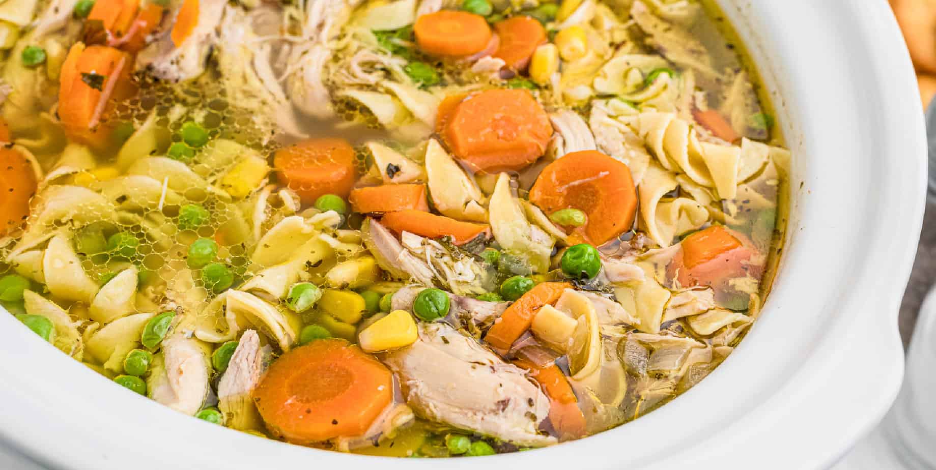 Wide view of inside a slow cooker close up full of chicken noodle soup with big chicken pieces, egg noodles, carrots, peas and corn.