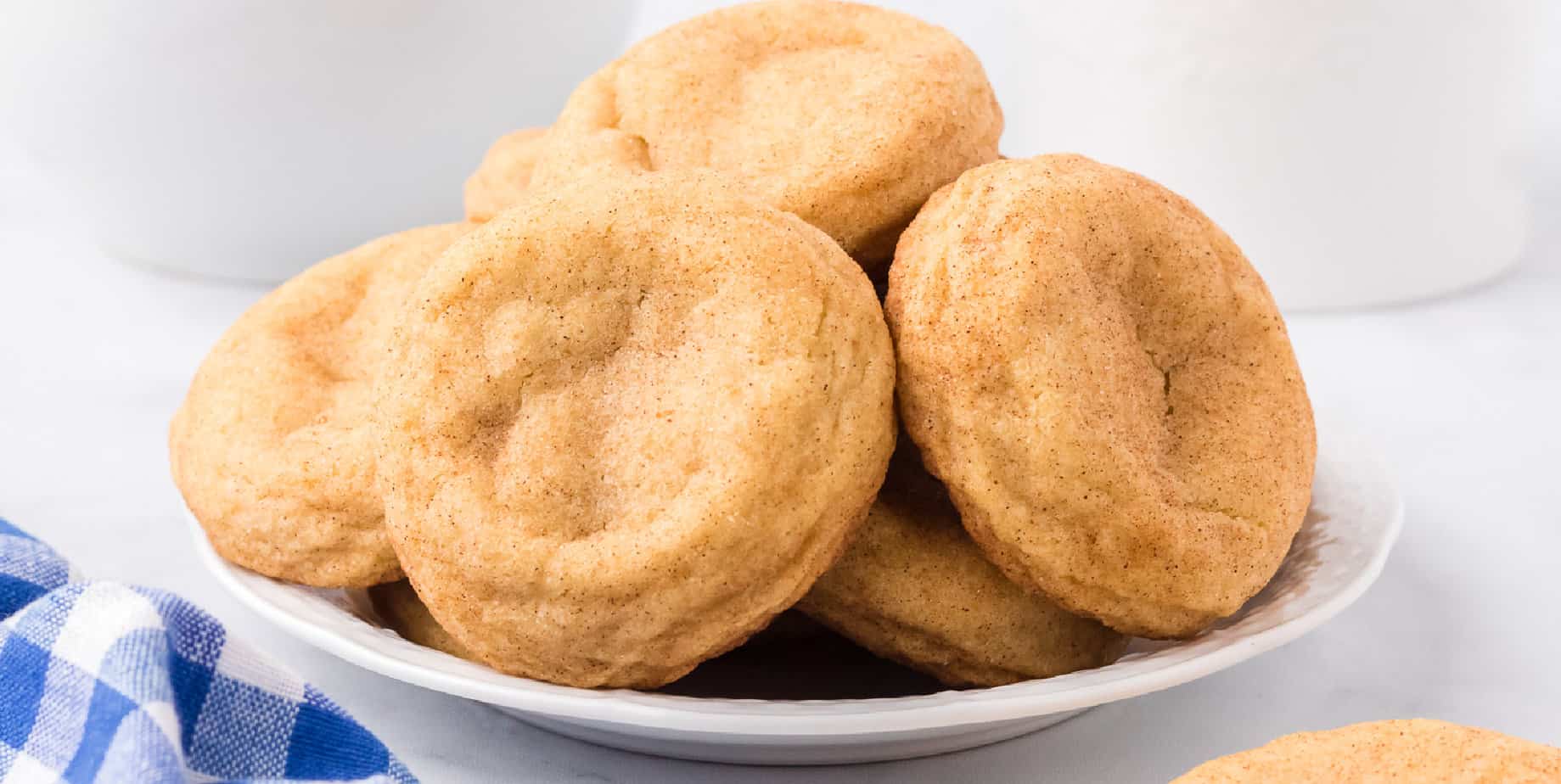 Snickerdoodles piled high on a plate from the side with a napkin and pitcher of milk nearby on the couneter.