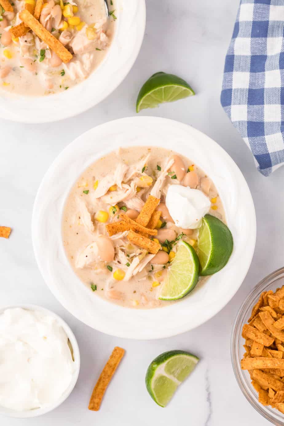 Bowl of creamy chicken tortilla soup from overhead on a counter topped with avocado, tortilla strips, live and sour cream next to a second bowl of soup and more toppings.