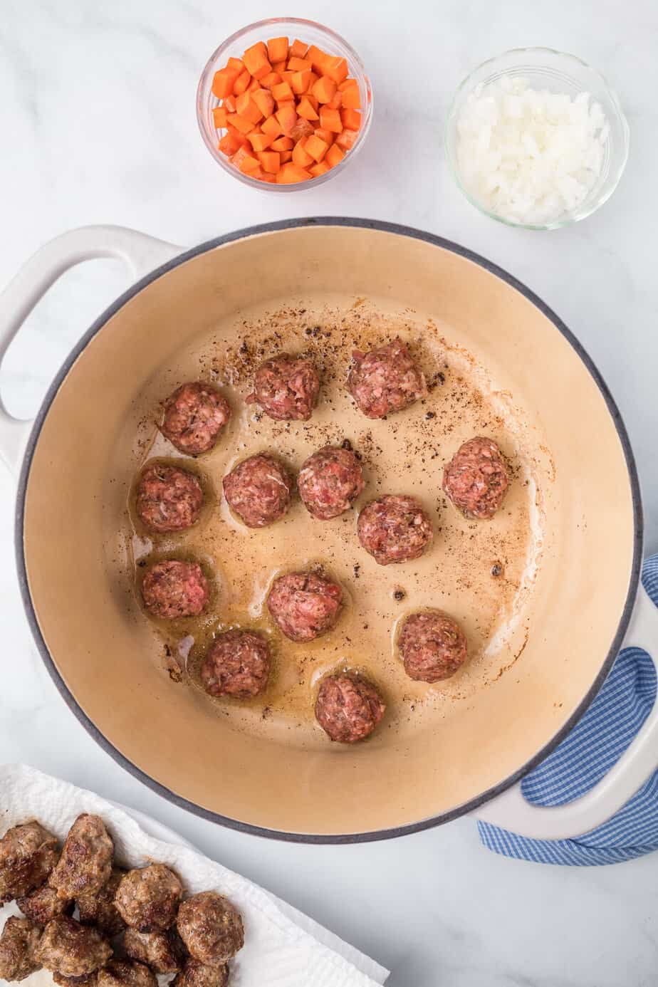 Browning meatballs in a pot from overhead on a counter with more cooked meatballs on a paper towel on a plate nearby.
