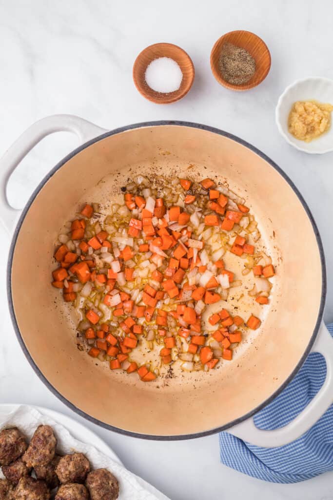 Cooking carrots and onions in a pot from overhead with meatballs and more soup ingredients on the counter nearby for Italian Wedding soup.
