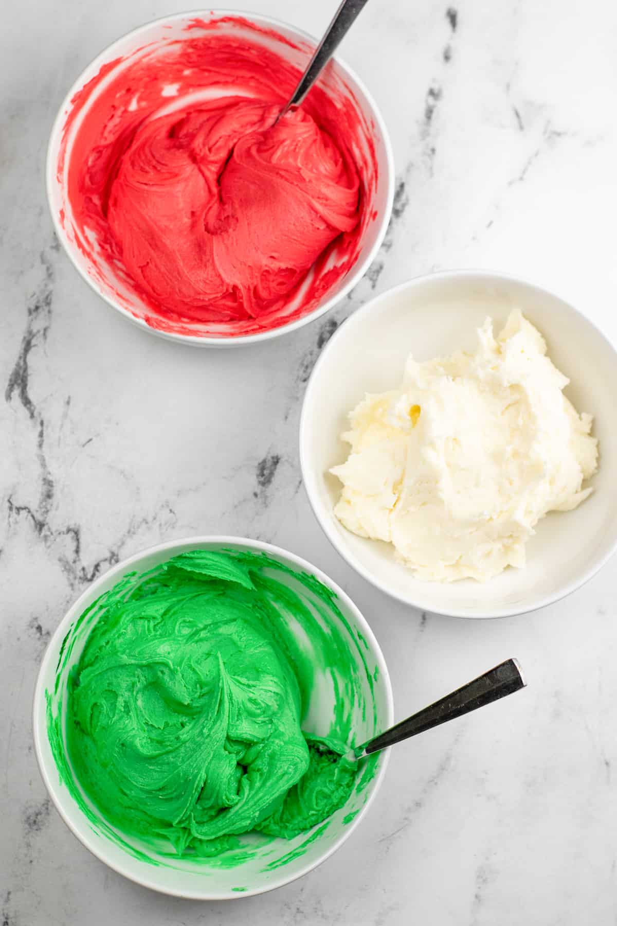 Red white and green cream cheese mint dough each in their own bowl on a counter from overhead with spoons in the red and green dough.