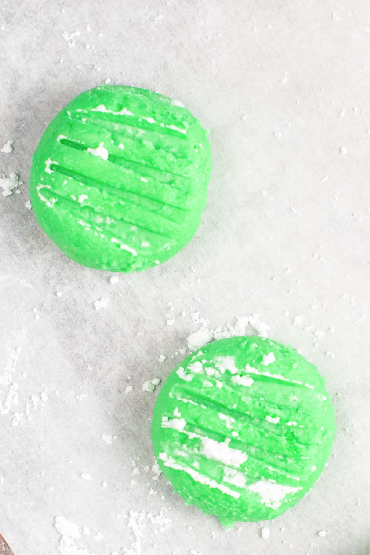 Two green cream cheese mints pressed flat with a fork to make grooves up close on parchment.
