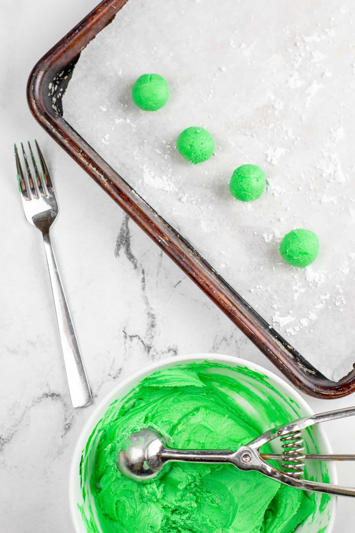 Scooping green cream cheese mint dough from a bowl with a cookie scoop into small balls on a baking sheet lined with parchment paper from above.