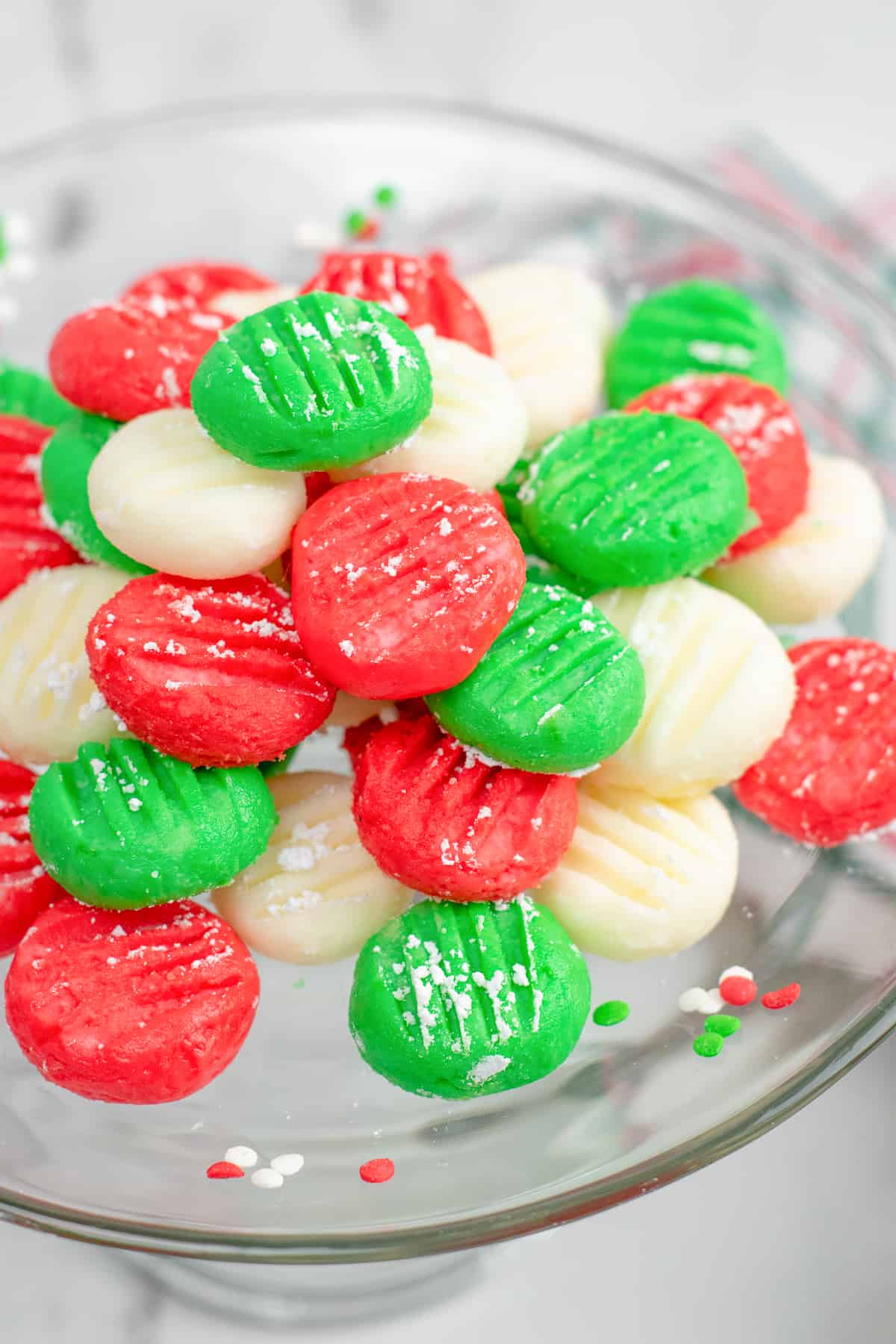 Red, green and white cream cheese mints stacked high on a clear plate from the side on a counter.